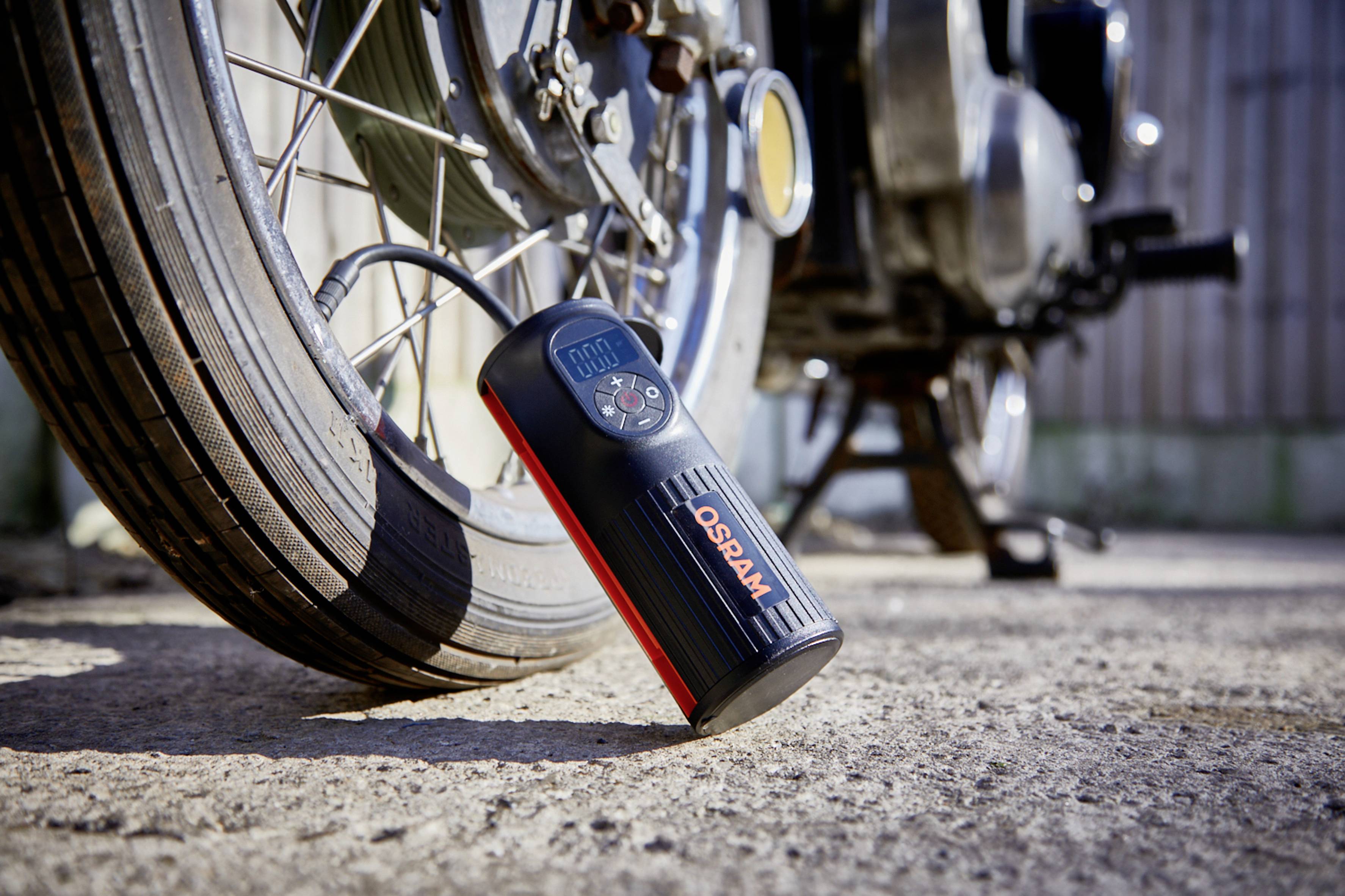 Přenosný kompresor stojí vedle motocyklové pneumatiky a nafukuje ji. Display ukazuje 0,00 psi. Slunce svítí na scénu.