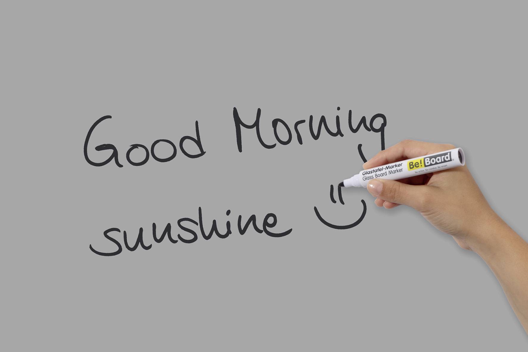 Ruka píše propiskou na šedém pozadí: 'Good Morning sunshine' a namaluje smějící se obličej.