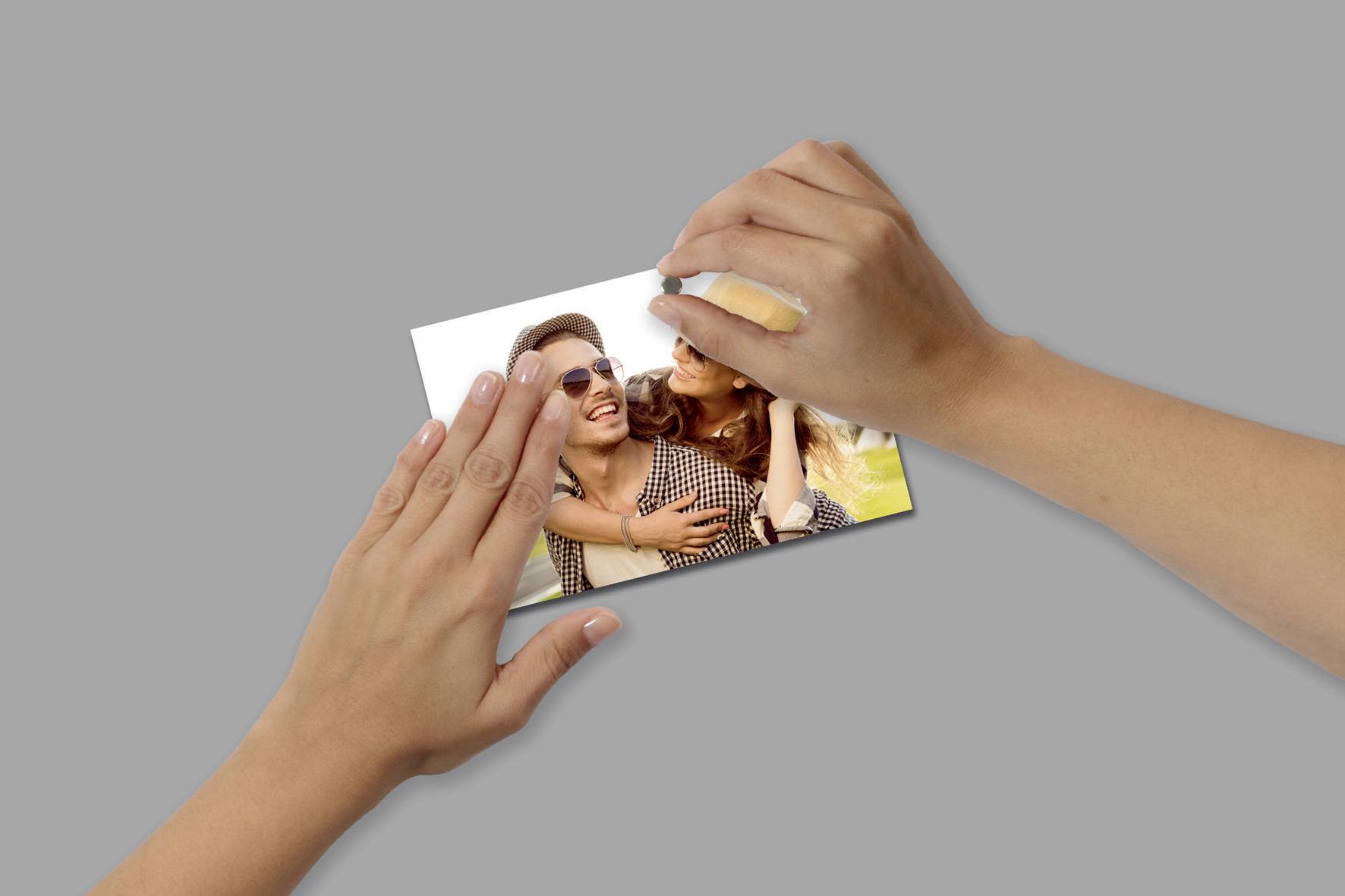 Dvě ruce drží fotografii usmívajícího se páru, který se objímá. Muž má sluneční brýle. Pozadí je rozmazané.