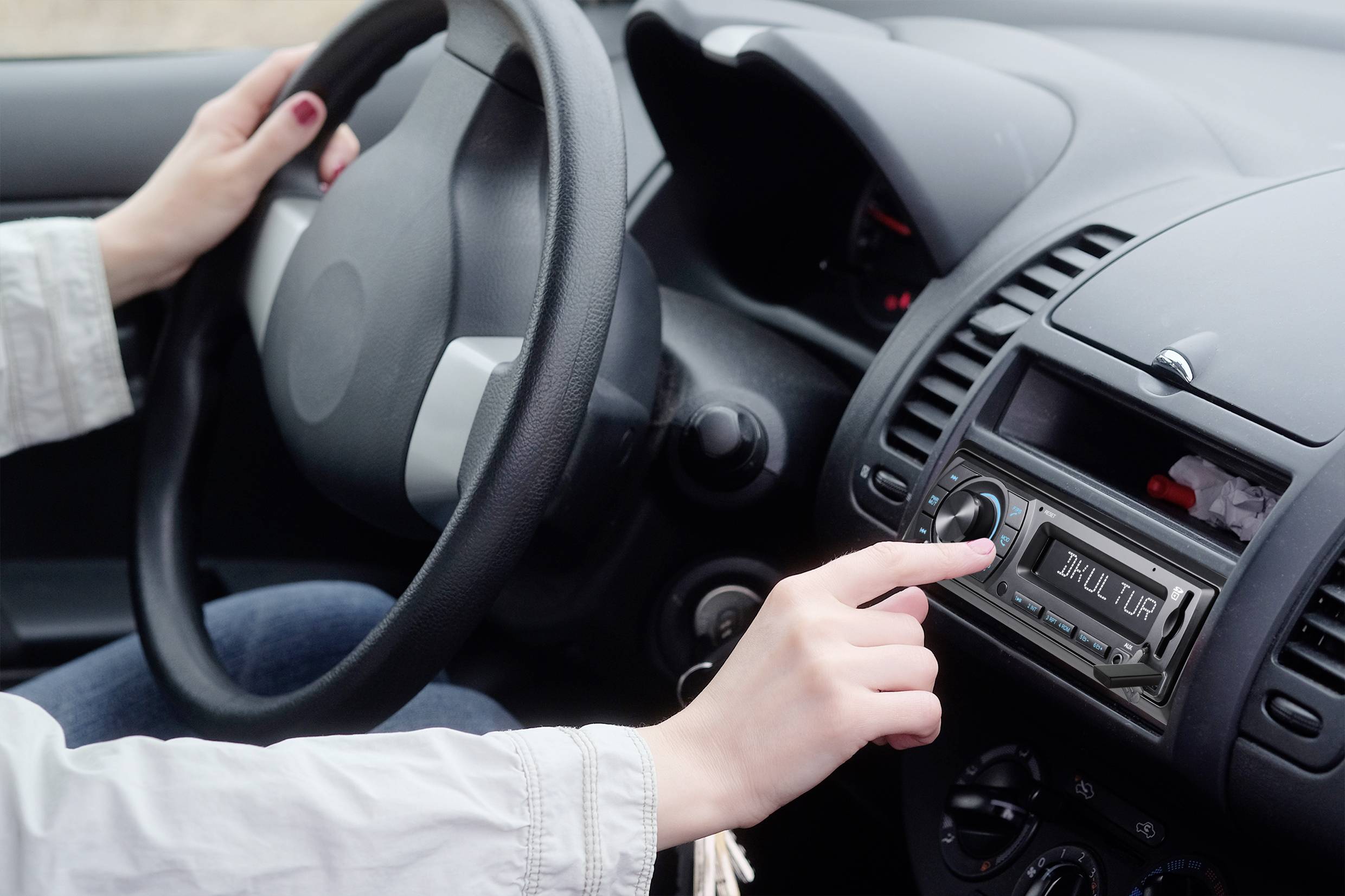 Řidič ovládá automobil a nastavuje rádio na přístrojové desce. Zaměření je na ruku na rádiu a volantu.
