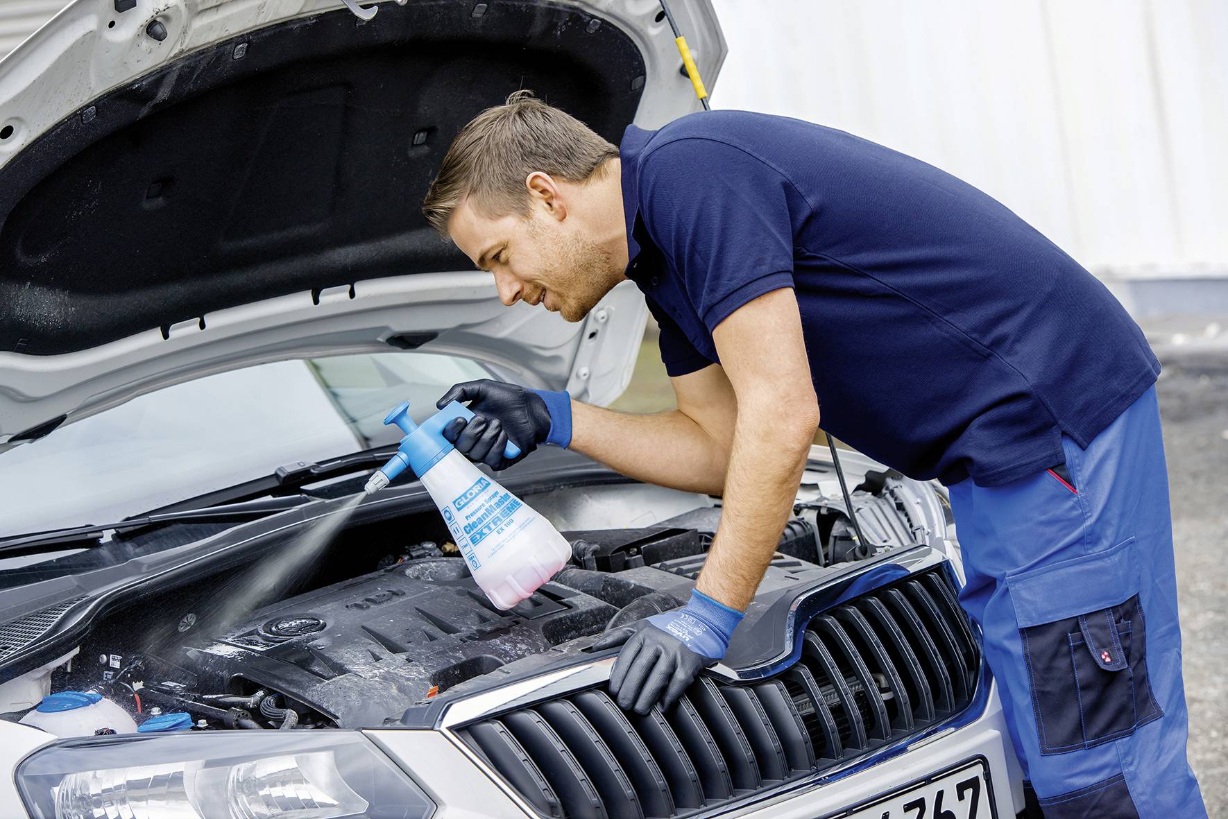 Mechanik čistí motorový prostor automobilu sprejem. Střecha vozu je otevřená a muž má na sobě pracovní rukavice.