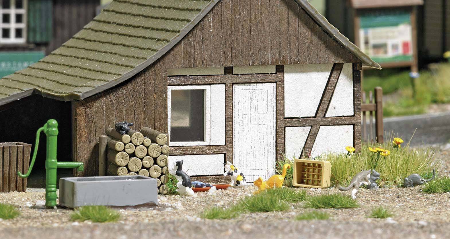 Malý modelový dům s hrázděným zdivem, obklopený hromadami dřeva, ruční pumpou a miniaturními zvířaty na dvoře.