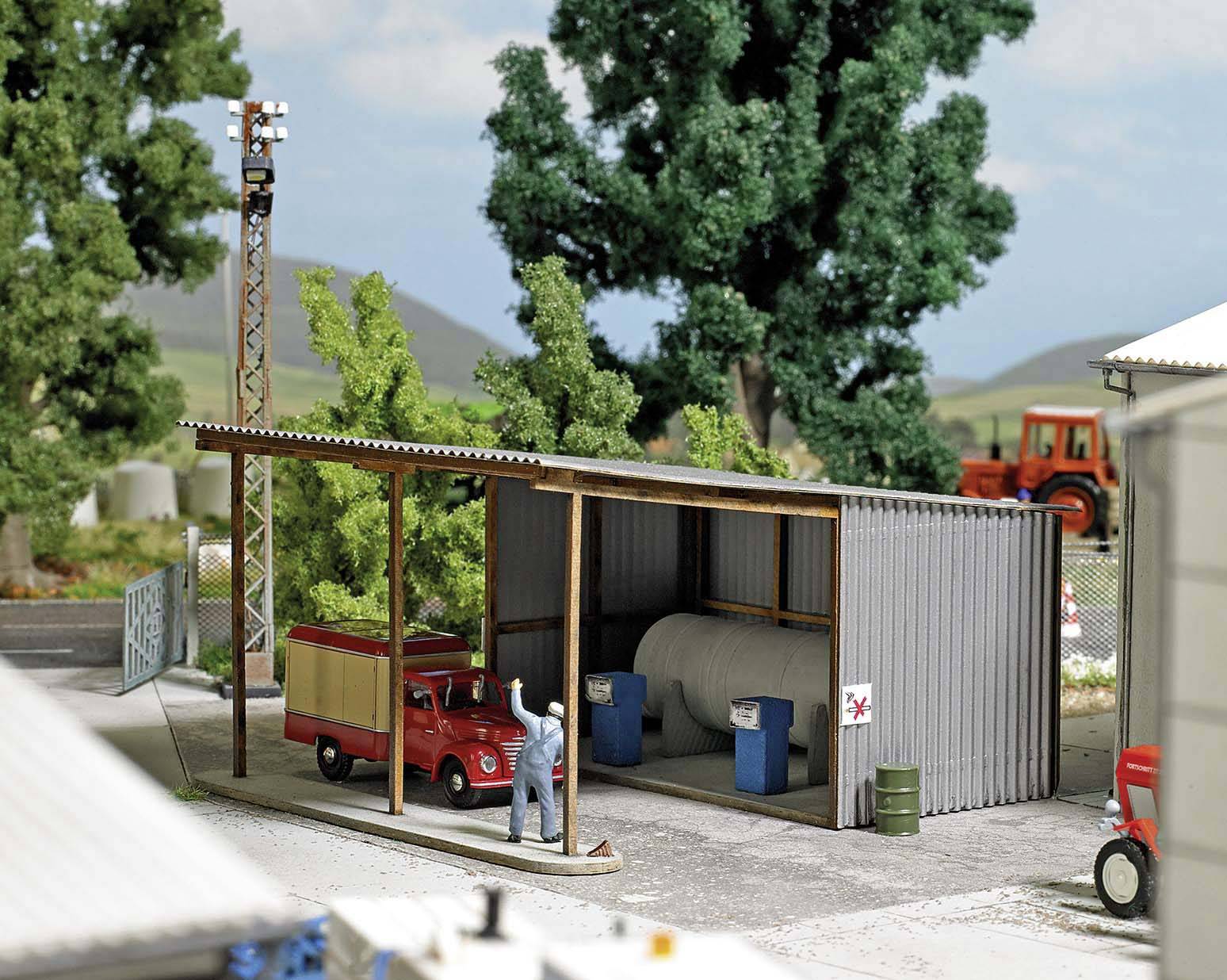 Model zemědělské budovy s miniaturní figurkou, červeným dodávkovým vozem a nádržemi. Stromy a traktor v pozadí.