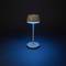 Moderní stolní lampa s jednoduchým designem svítí modrým světlem a stojí na tmavém pozadí.