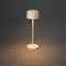 Minimalistická stolní lampa s kulatým bílým stínidlem a úzkým podstavcem osvětluje teplým světlem tmavý povrch.