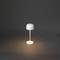 Bílá stolní lampa s kulatým stínítkem a štíhlou základnou, tlumeně svítící v tmavém prostoru. Minimalistický design.