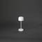 Bílá stolní lampa s moderním designem na tmavém pozadí. Válcovitý stínidlo umístěné na štíhlé podstavě.