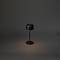 Minimalistická černá stolní lampa na tmavém pozadí, která vrhá jemný kužel světla na stůl.