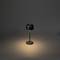 Černá stolní lampa svítí na tmavém pozadí. Má minimalistický styl a vytváří teplý světelný kužel.