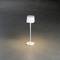 Štíhlá bílá stolní lampa svítí na tmavém povrchu. Lampa má minimalistický, moderní styl.