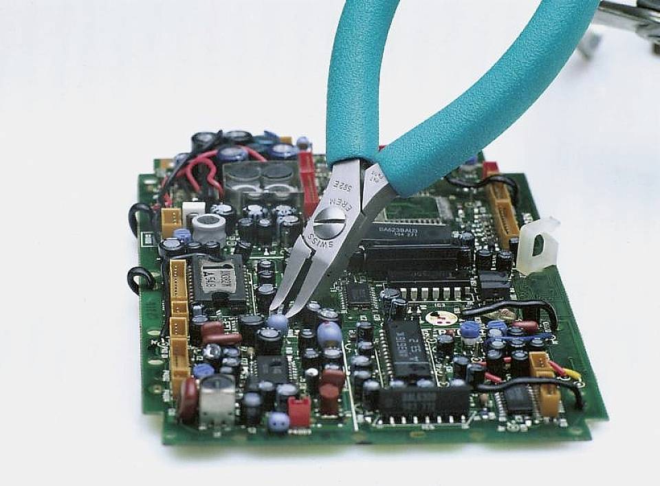 Kleště opracovávají elektrickou desku plošných spojů, která zobrazuje různé elektronické komponenty.