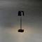 Černá stolní lampa s minimalistickým designem jemně osvětluje okolní šedou podlahu. Stínidlo lampy má tvar válce.