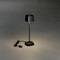 Černá stolní lampa s úzkým stojanem a čtvercovým stínidlem stojí na šedé podlaze. Kabel se zástrčkou leží vedle ní.