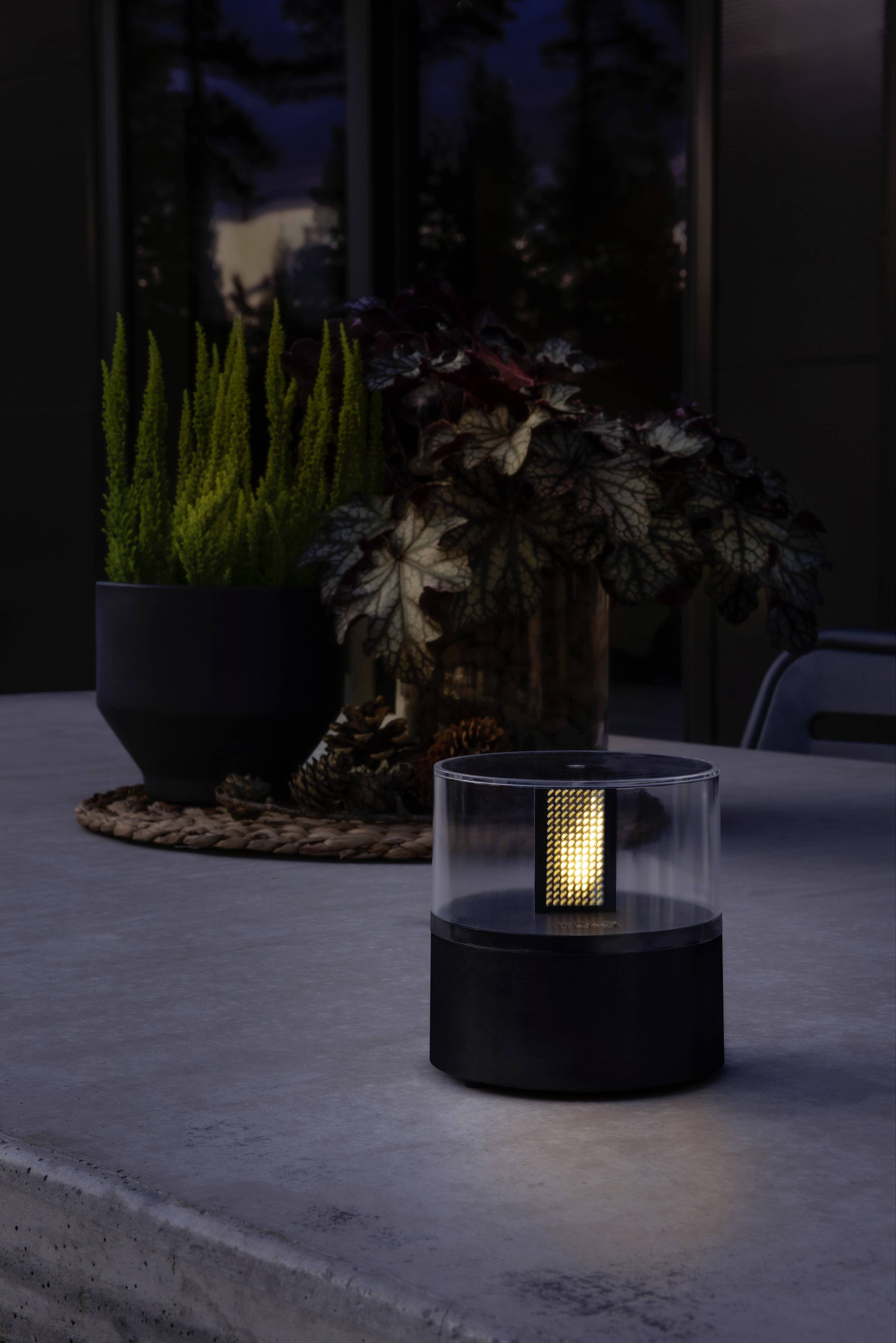 Malá, moderní stolní lampa s teplým světlem stojí na šedém stole. V pozadí jsou rostliny v tmavých květináčích.