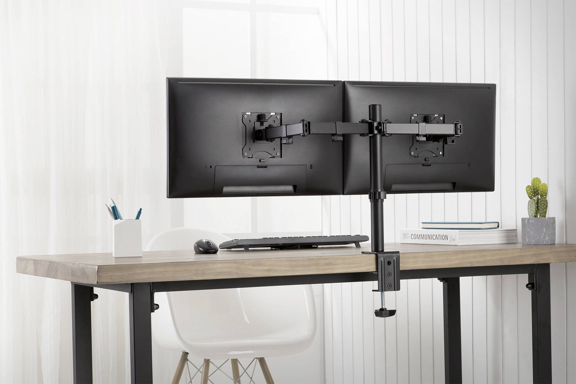 Dva monitory na pracovním stole s držákem pro dual monitor; v pozadí okna, vedle křeslo a dekorace.