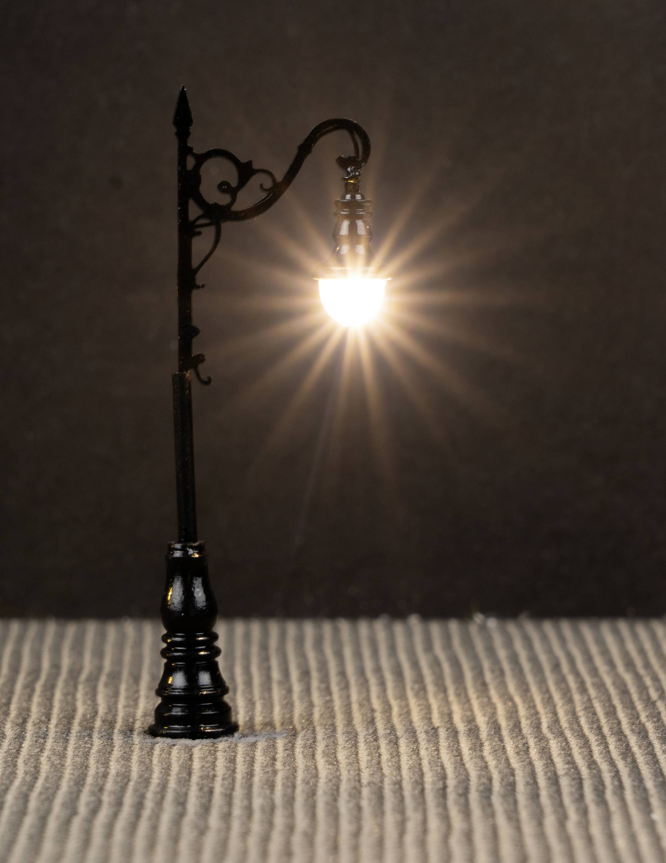 Malá pouliční lampa svítí jasně, vyzařuje teplé světlo na žebrovanou zem v tmavém pozadí.