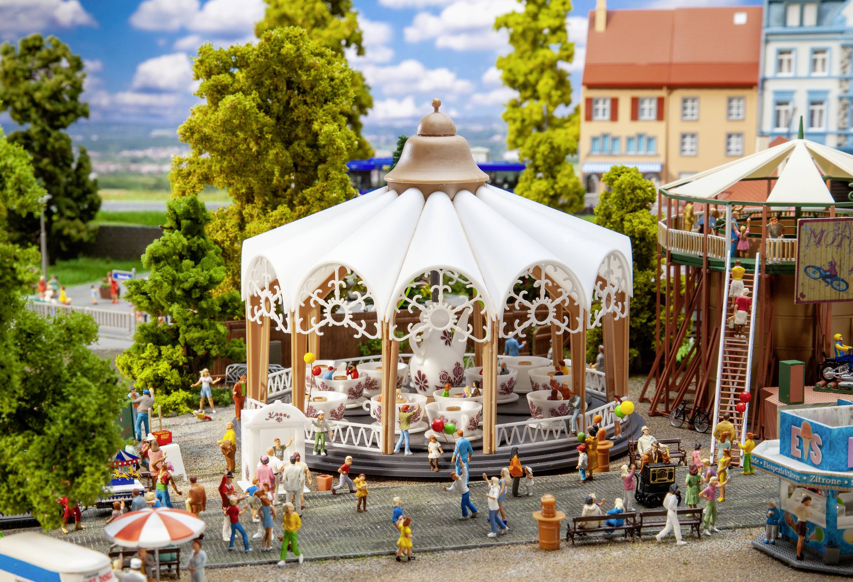 Miniaturní svět s pavilonem kolotoče, obklopený figurkami a stromy v městském parku za slunečného počasí. Děti a dospělí zde společně komunikují.