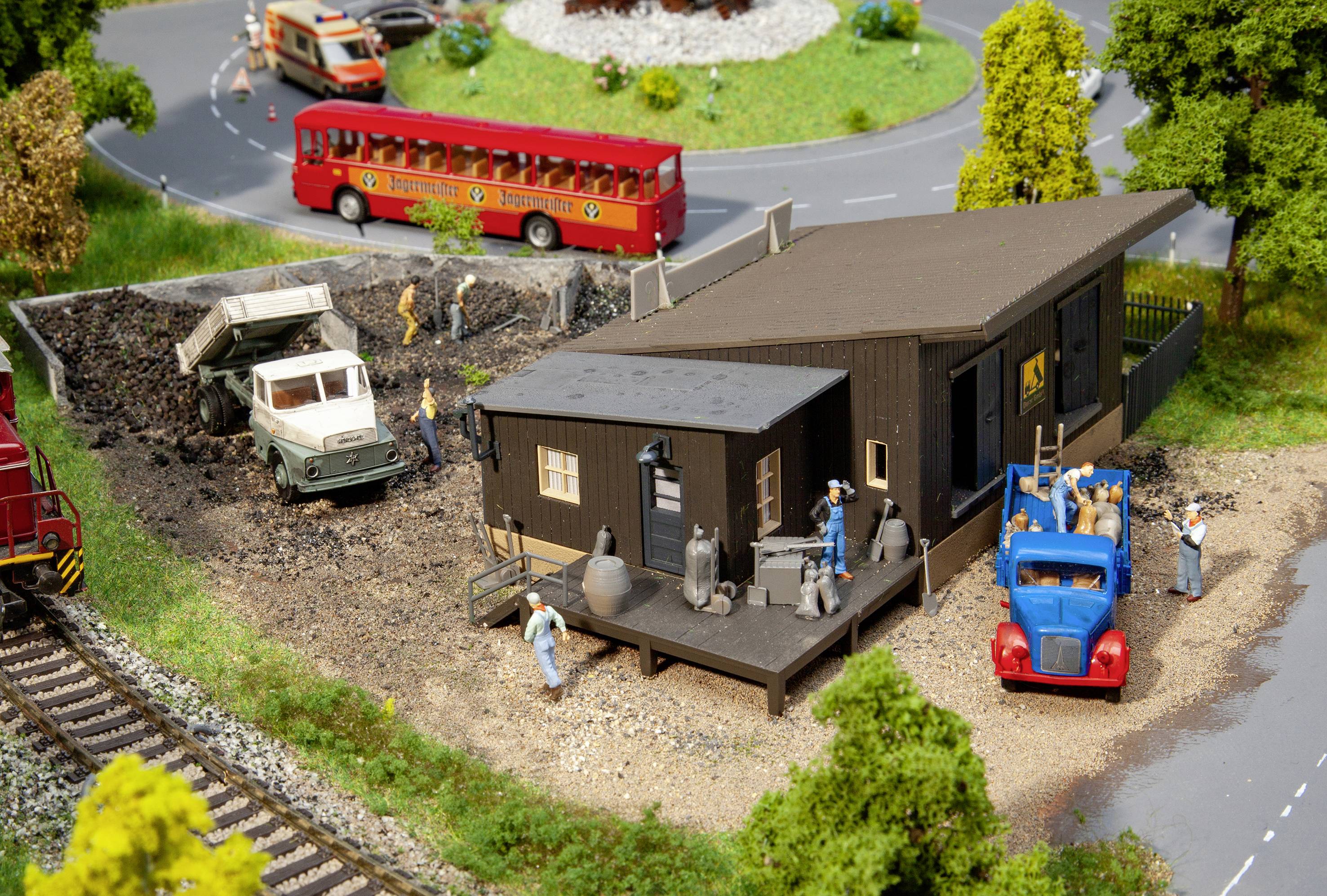 Diorama zobrazuje staveniště vedle silnice s nákladním automobilem, dělníky a červeným autobusem v pozadí.