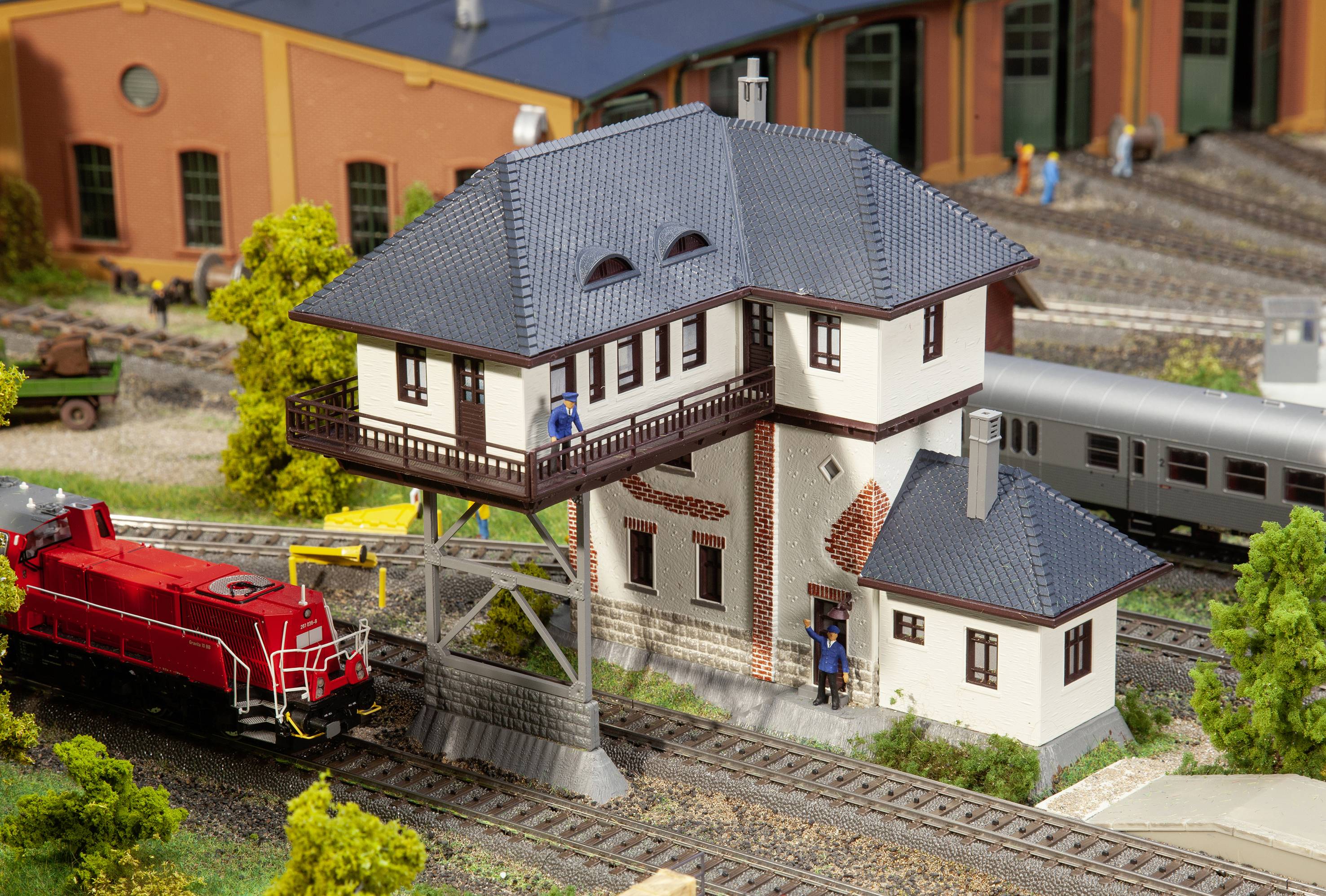 Miniaturní model zobrazuje železniční depo s červeným vlakem, bílou budovou a několika figurkami.