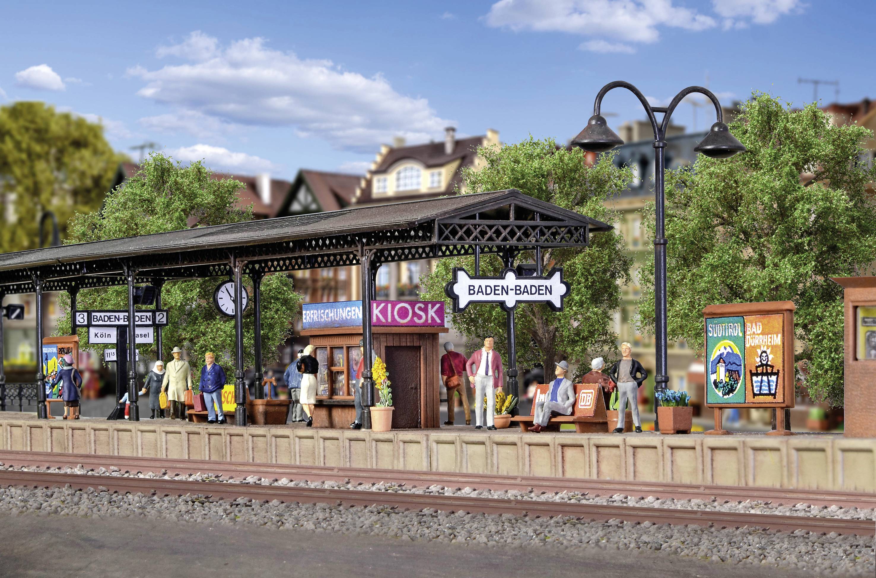 Miniaturní model nádraží s mnoha figurkami a cedulemi s nápisem 'Baden-Baden', obklopený budovami a stromy.