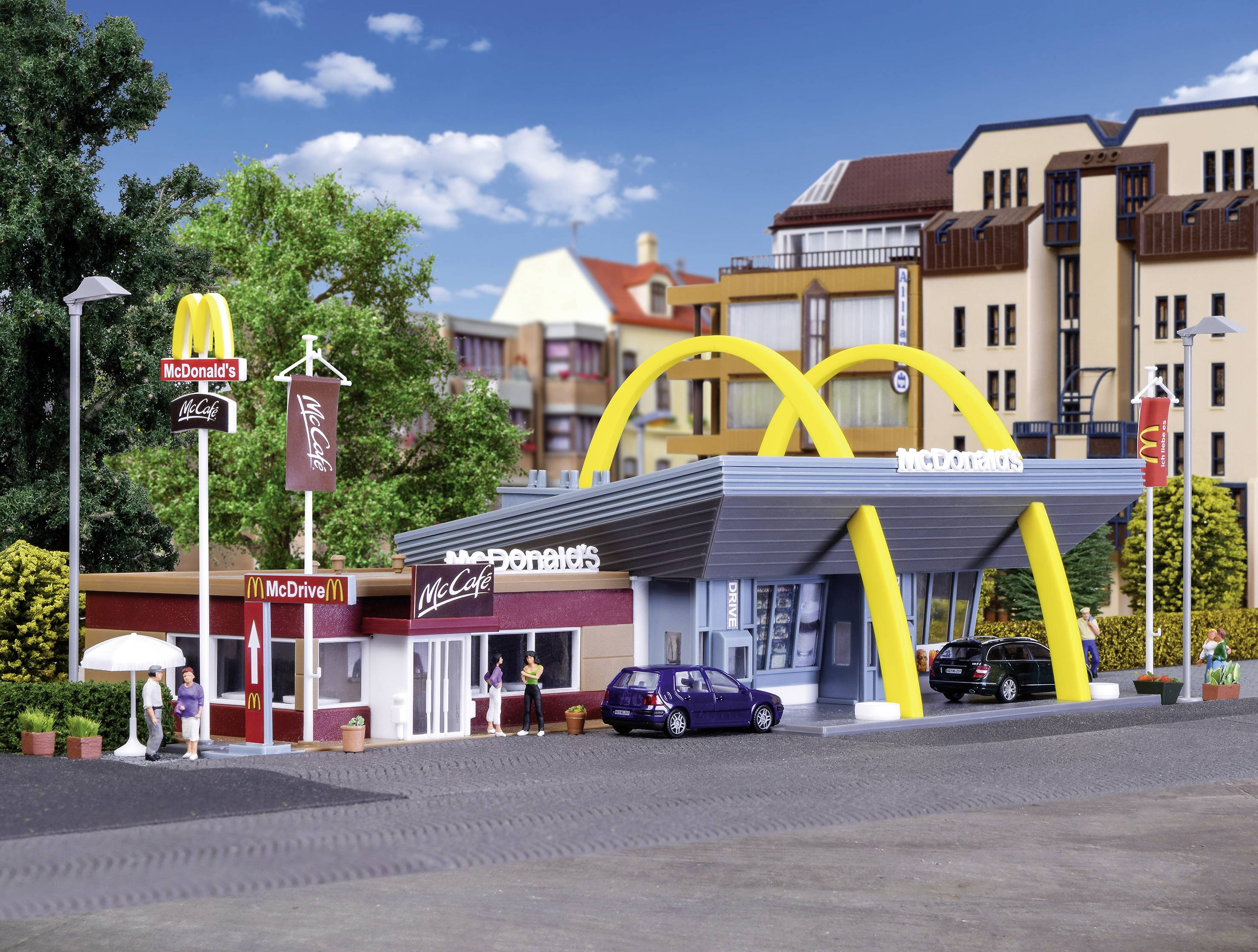 McDonald's restaurace se žlutými oblouky, kde lidé sedí venku. V pozadí moderní budovy a modrá obloha.