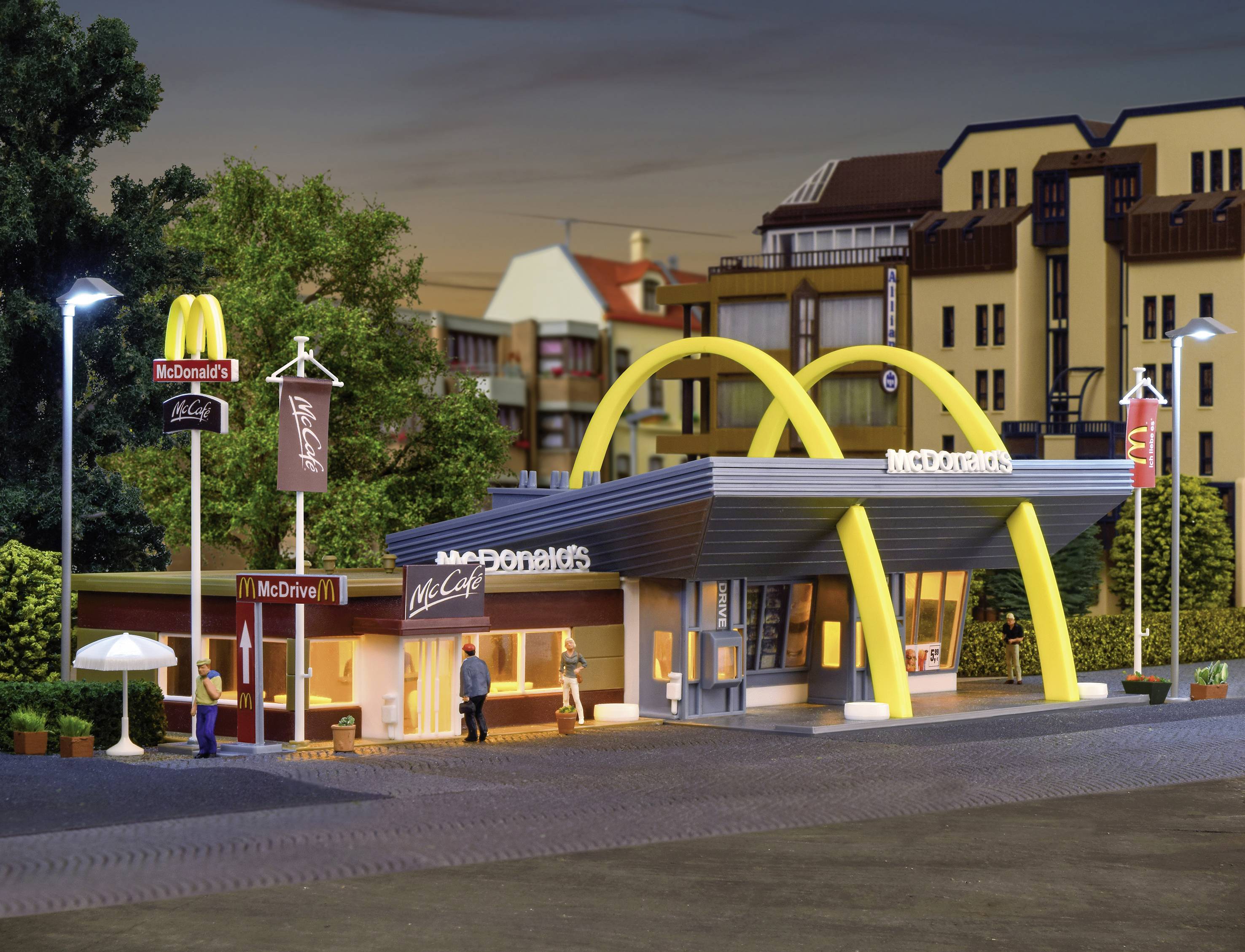 Model McDonaldova restaurace se žlutými oblouky, obklopený stromy a pouličními lampami, zobrazuje hosty před vstupem.