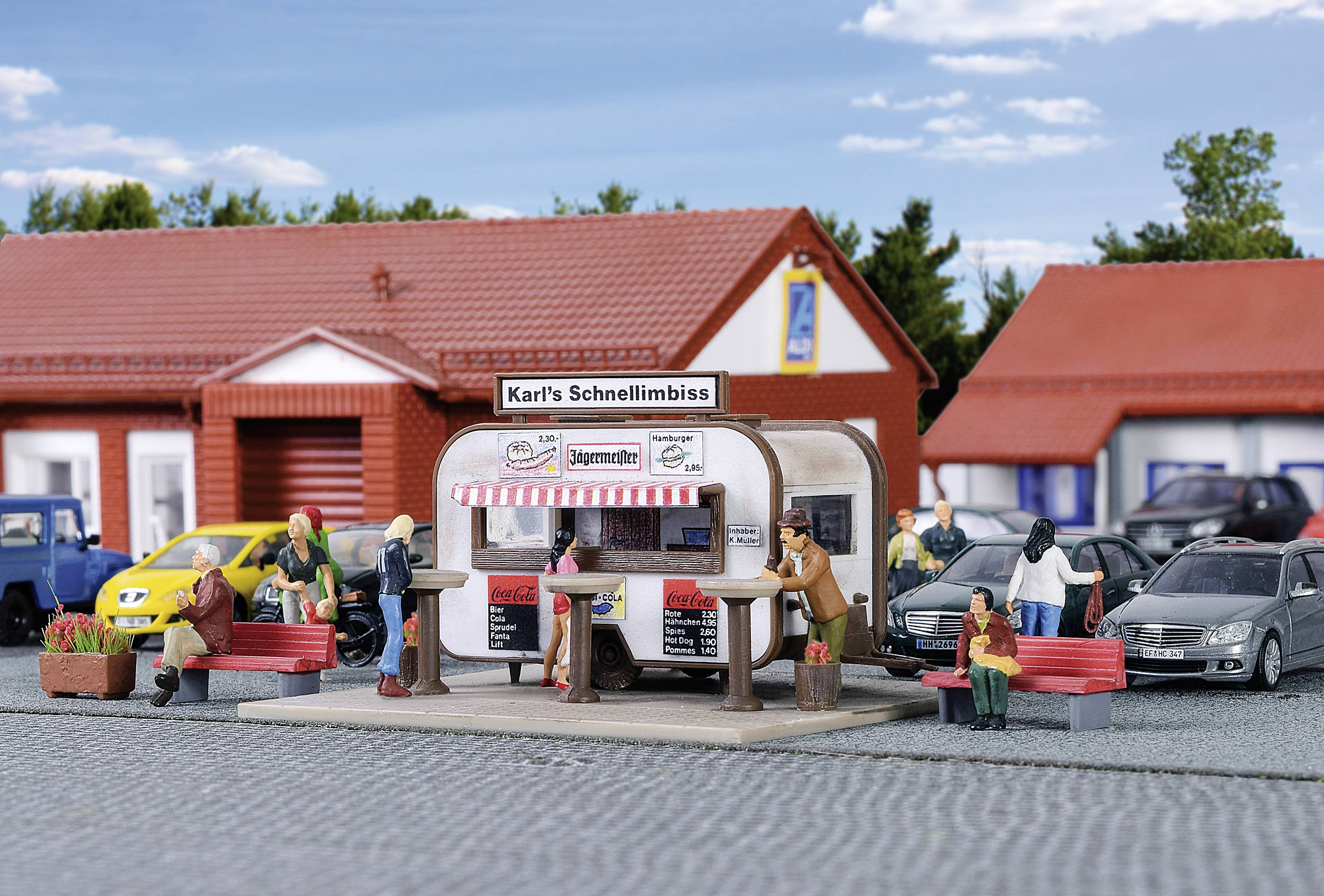 Miniaturní znázornění stánku s občerstvením, kde lidé sedí u stolů a jedí. V pozadí jsou auta a budovy.