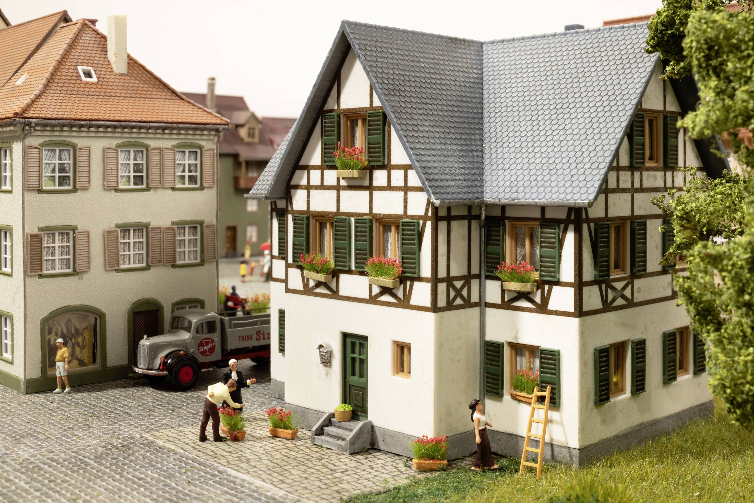 Miniatura hrázděného domu s kvetoucími truhlíky, obklopená figurkami v vesnici. Dodávka je zaparkovaná vedle něj.