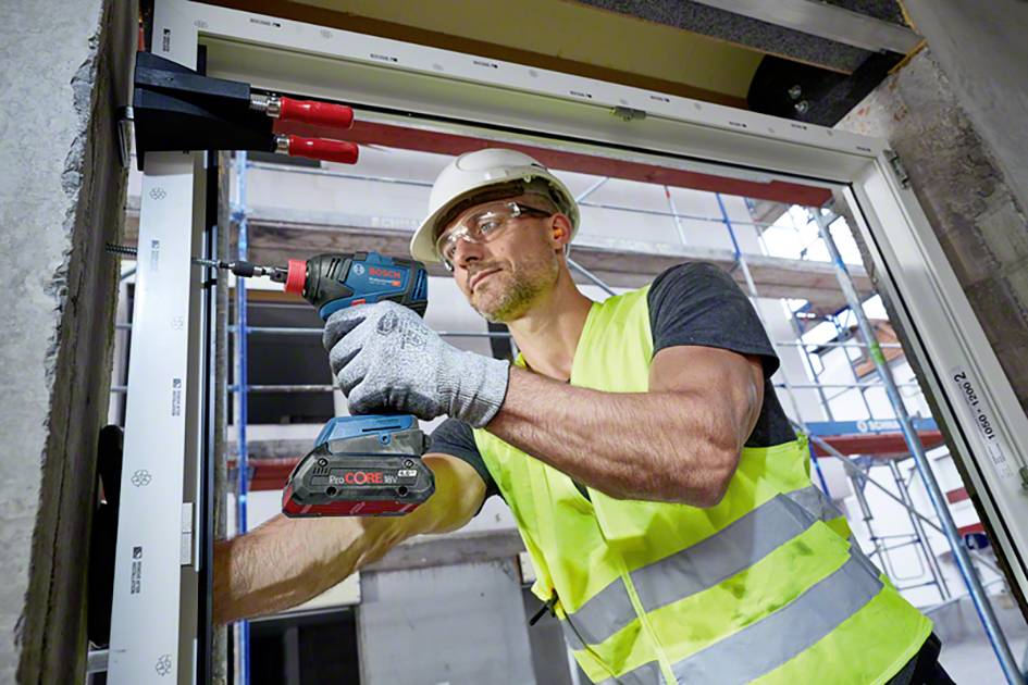 Dělník v ochranném oděvu a helmě upevňuje okno pomocí vrtačky v budově, v pozadí je vidět lešení.