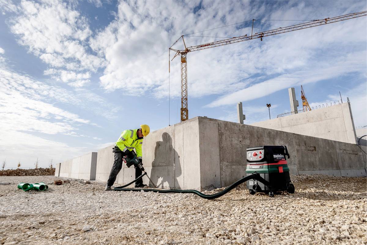 Dělník v ochranném oděvu pracuje s těžkou technikou na staveništi. V pozadí je vidět jeřáb.
