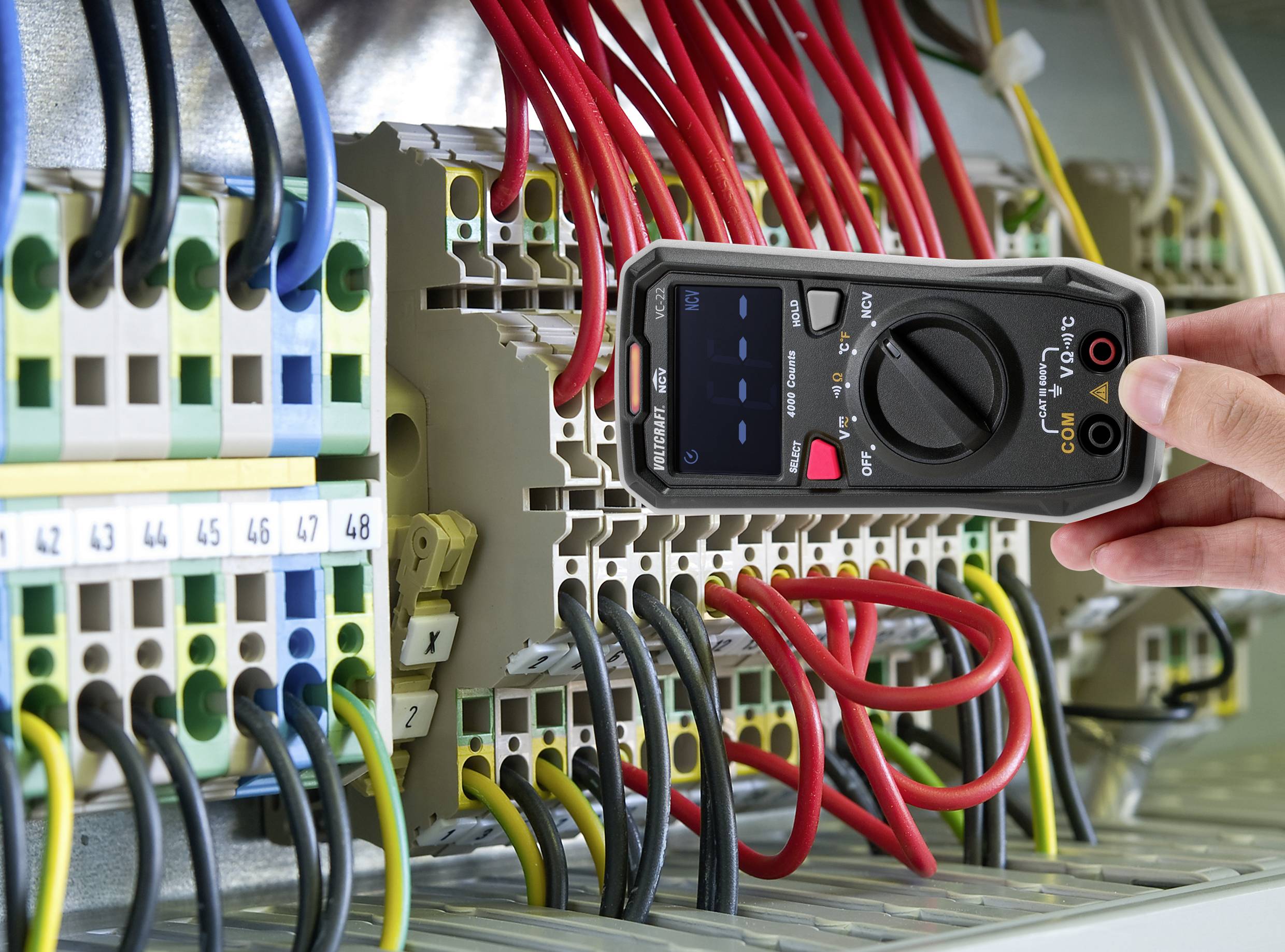 Osoba používá multimetr k měření napětí v elektrickém rozvaděči. Barevné kabely jsou připojeny.