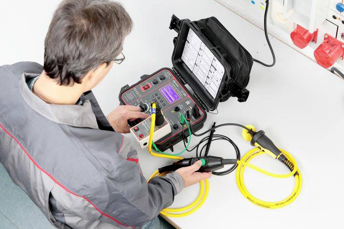 Osoba obsluhuje elektrické testovací zařízení u stolu. Přístroj má žluté kabely a displej zobrazující naměřené údaje.