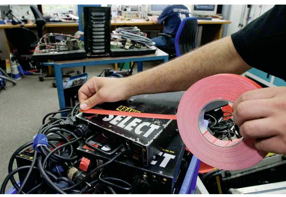 Osoba lepí červenou lepicí pásku s nápisem 'CABLE SELECT' na elektronické zařízení. V pozadí je pracoviště s elektronikou.