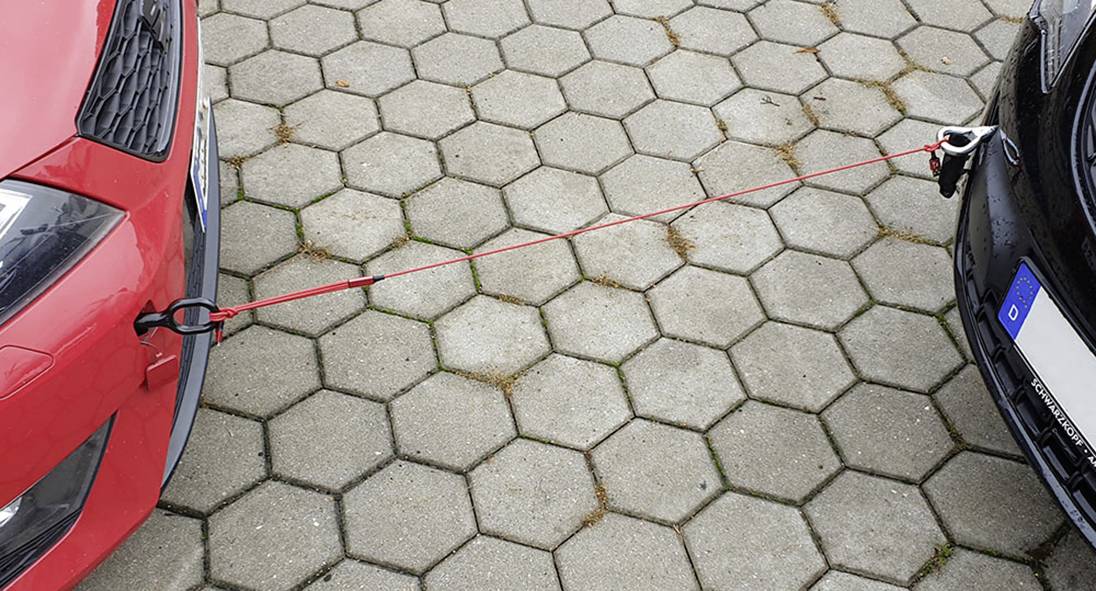 Červené a černé auto jsou propojeny červeným odtahovým lanem, které leží na dlážděném parkovišti.
