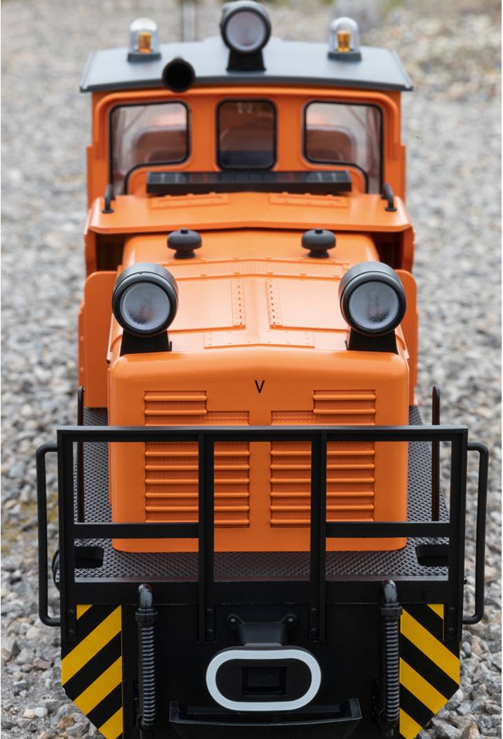 Malá oranžová lokomotiva na štěrkovém chodníku, fotografovaná čelně. Má dva velké světlomety a černý ochranný kryt vpředu.