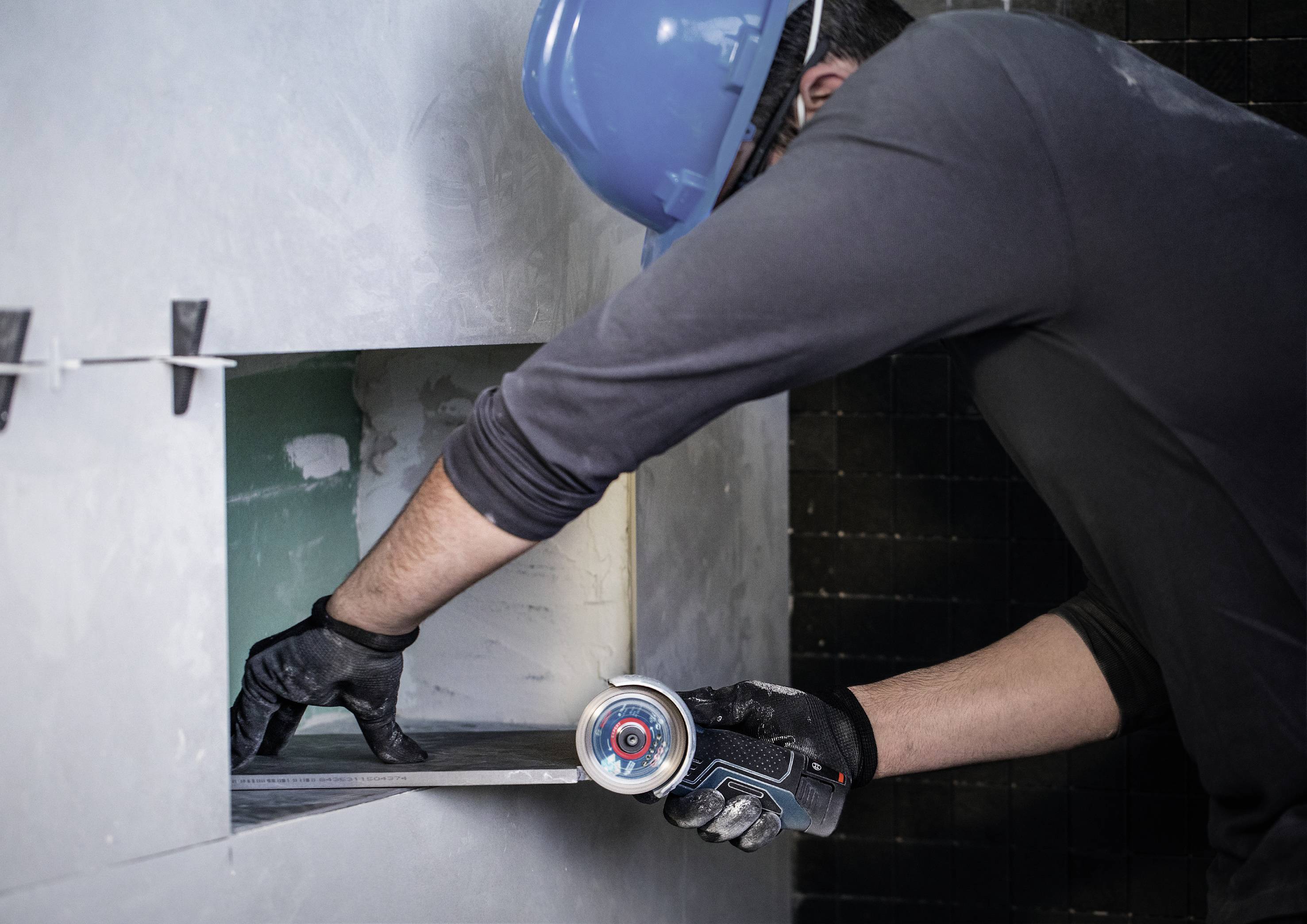 Osoba s modrým ochranným helmem a černými rukavicemi řeže úhlovou bruskou kovový díl v otvoru ve zdi.