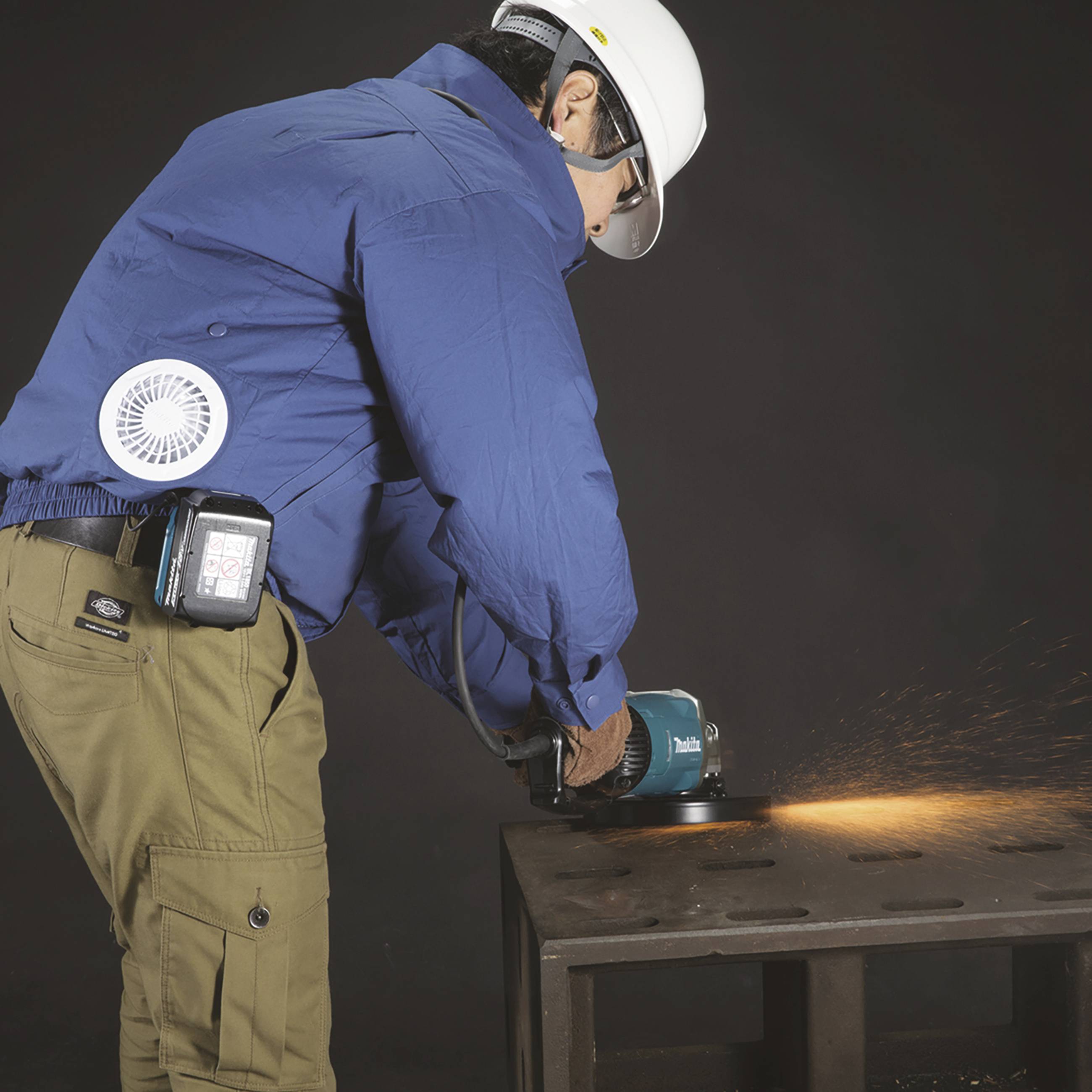 Dělník v ochranném oblečení brousí kov, přičemž kolem něj létají jiskry. Na oblečení má připevněnou větrací jednotku.