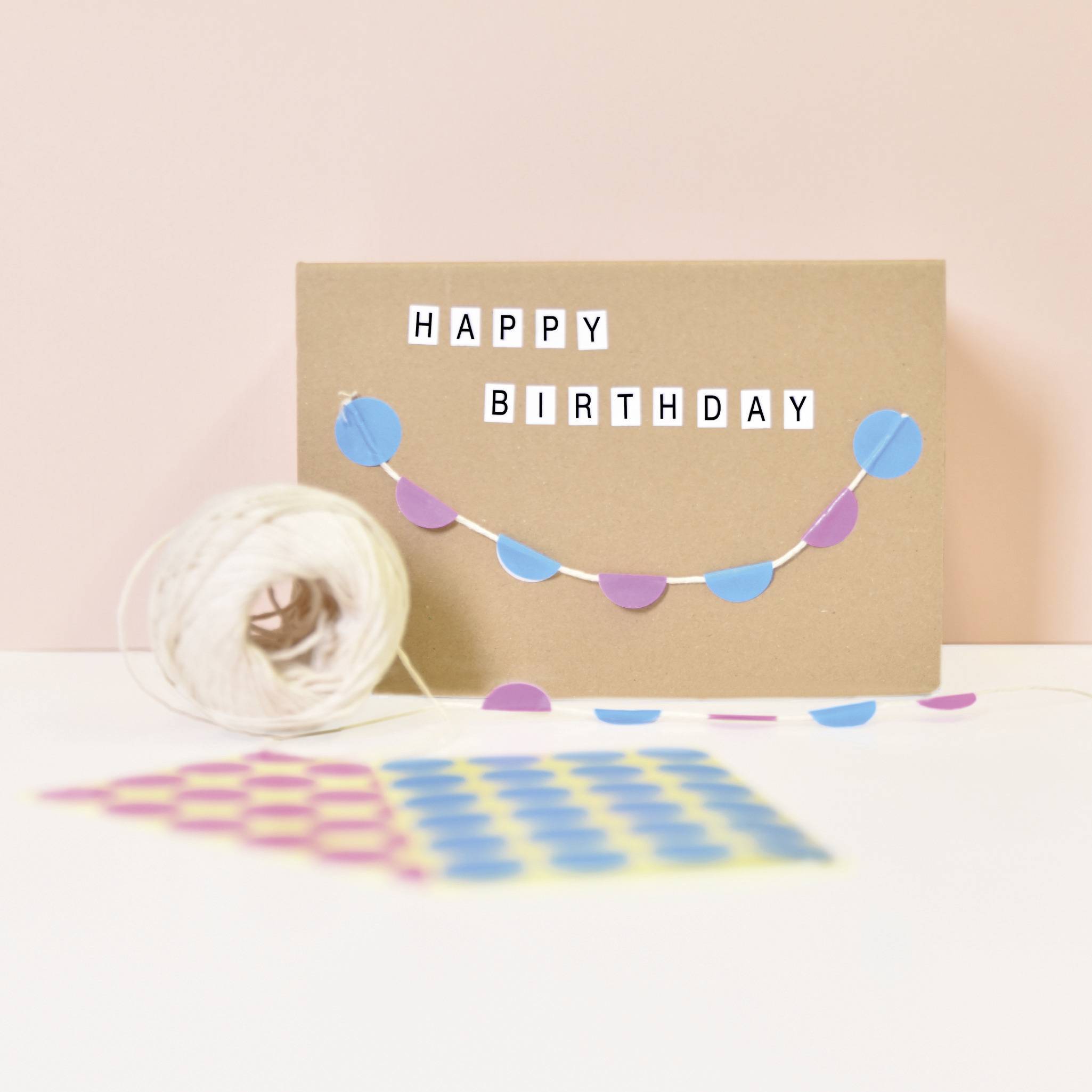 Narozeninová pohlednice s barevným praporkem a nápisem 'Happy Birthday' na hnědém pozadí, vedle leží klubko příze.