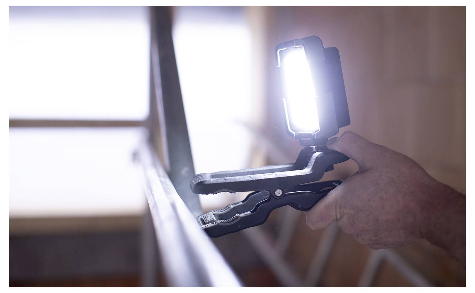 Osoba používající přenosnou LED pracovní lampu připevněnou na držák při práci v interiéru, se silným světlem zaměřeným na povrch.