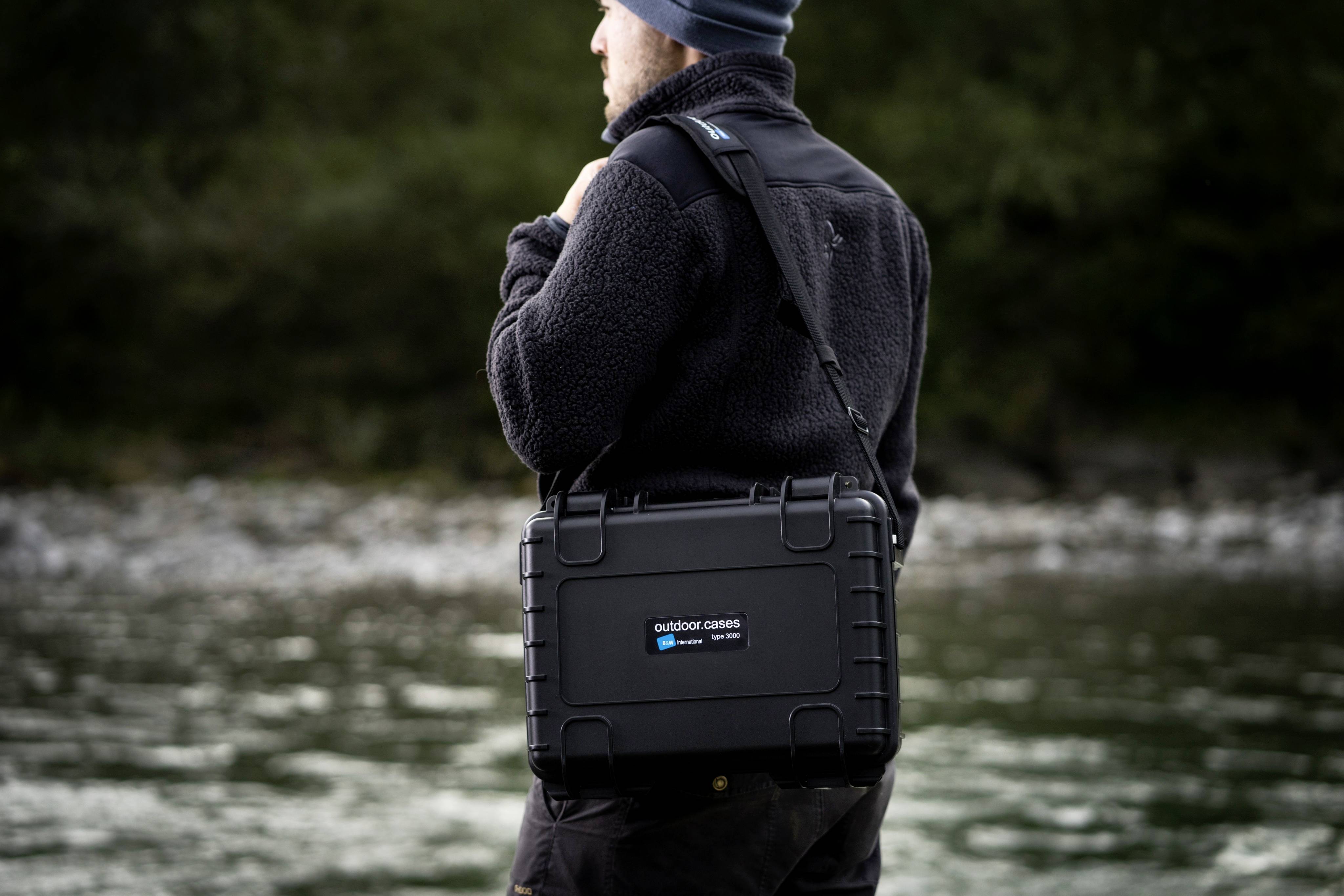 Osoba v černém outdoorovém oblečení nese robustní černý ochranný kufr s nápisem 'outdoor cases' na břehu řeky.