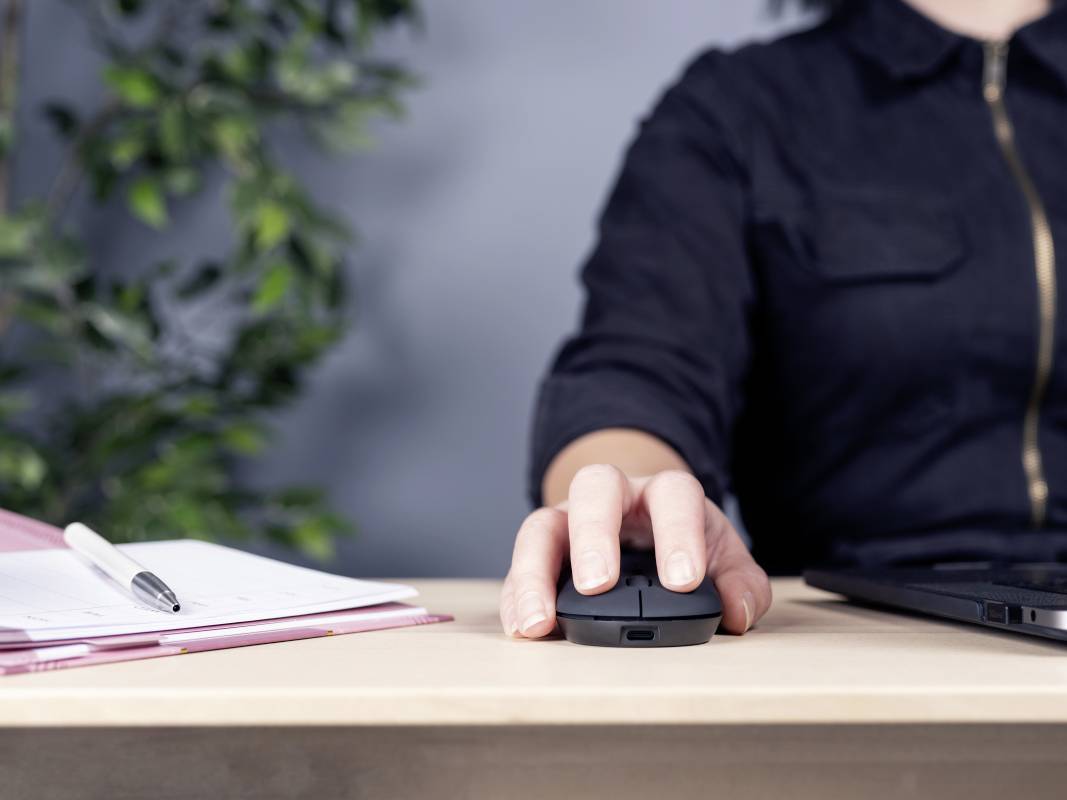 Osoba sedící u psacího stolu používá počítačovou myš. Vlevo jsou poznámkový blok a pero, vpravo laptop. V pozadí je rostlina.