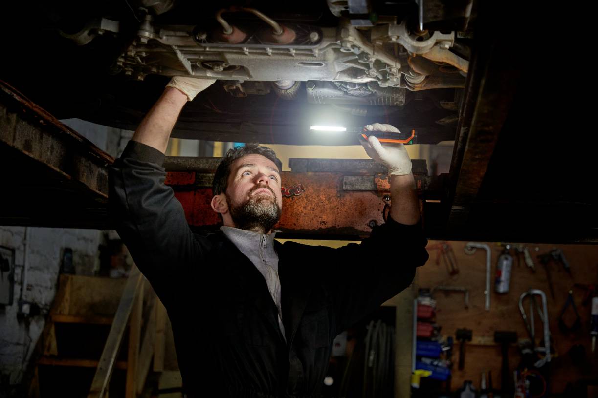 Mechanik pod autem v autodílně, soustředěně drží v ruce svítilnu a provádí opravy.