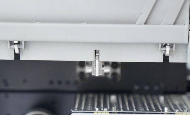 Detailní záběr průmyslového stroje s pohyblivou kovovou součástí nad děrovaným kovovým plechem.