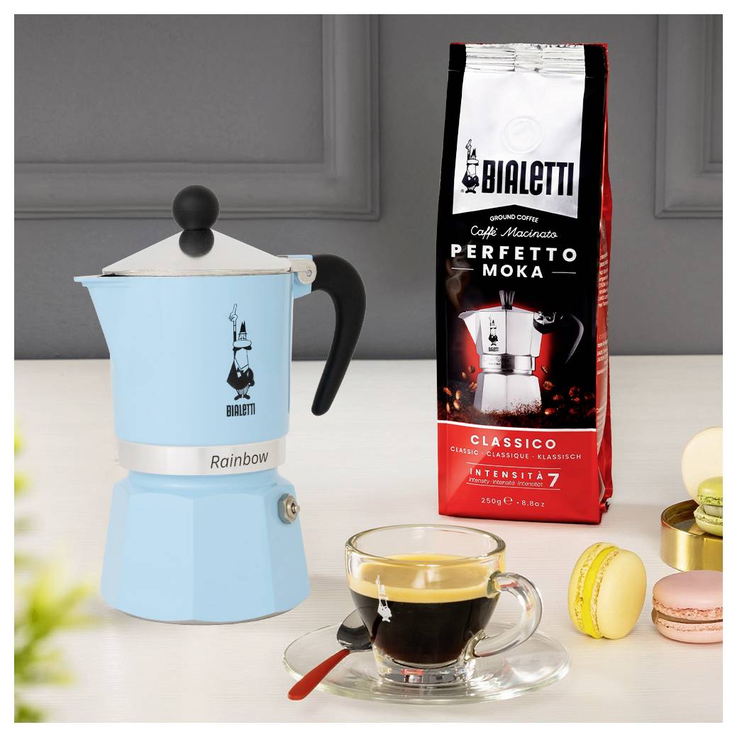 Modrá moka konvice, červený balíček moka kávy Bialetti Perfetto a espresso ve skleněné šálce jsou položeny na pultu před šedou stěnou.