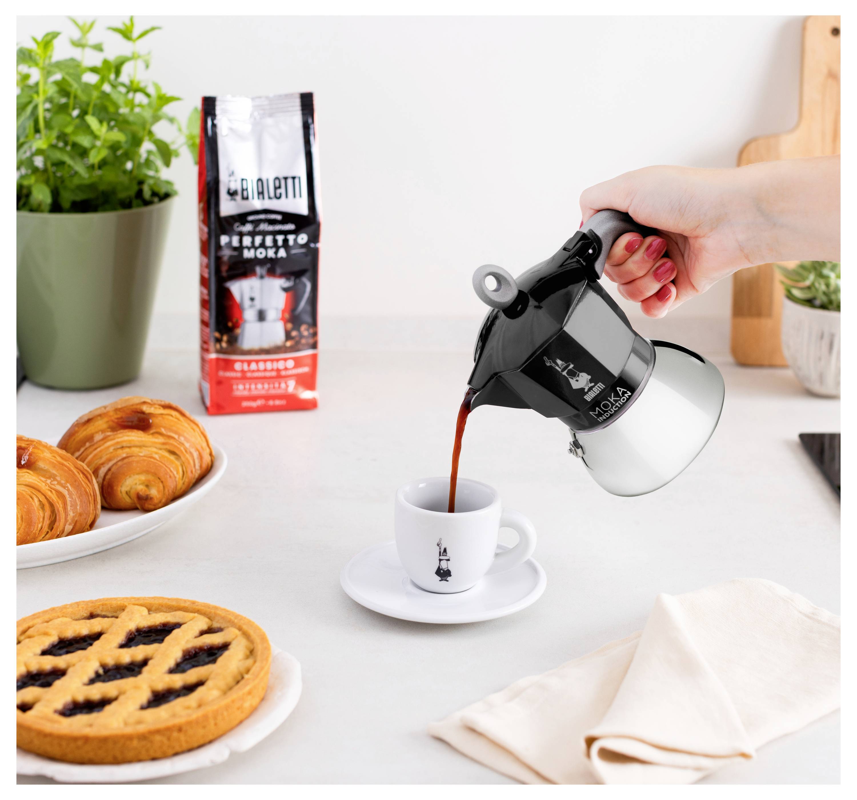 Člověk nalévá kávu z Moka konvičky do espresso šálku. Poblíž jsou croissanty, koláč a kávový sáček Bialetti na kuchyňské lince.