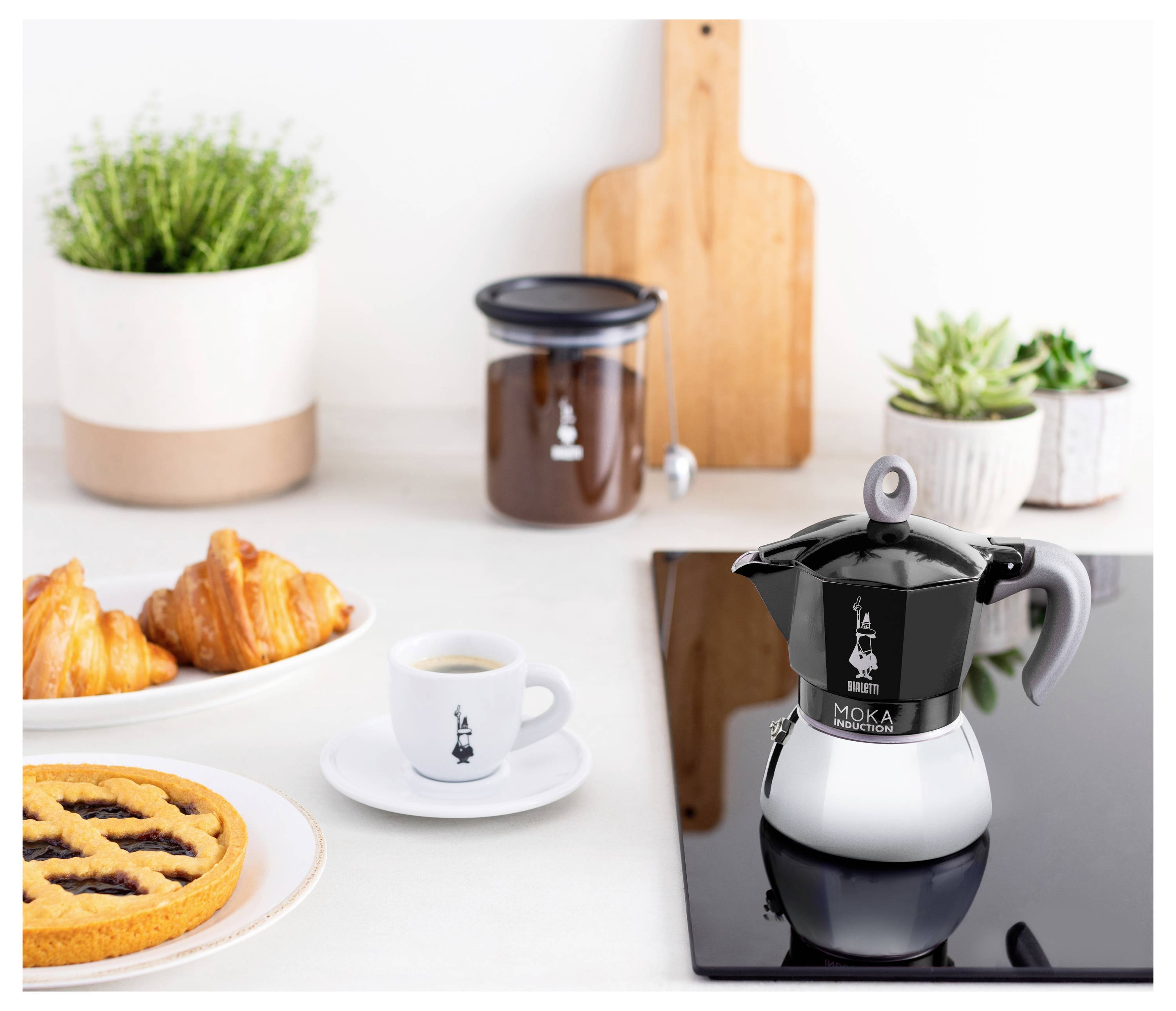 Moka konvice na kuchyňské lince spolu s šálkem kávy, croissanty, koláčem a sklenicí kávy, vytvářející útulné prostředí pro snídani.