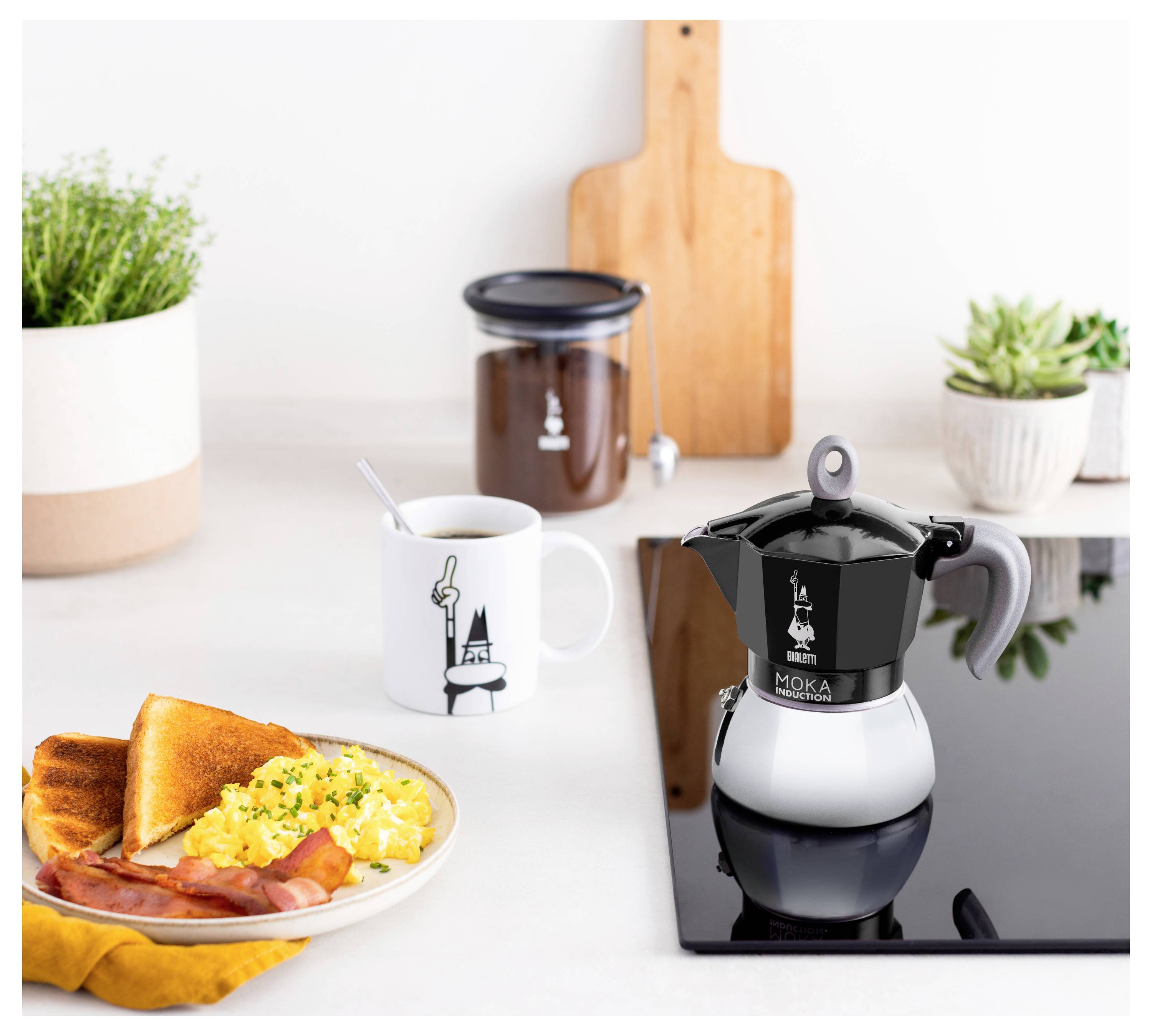 Kuchyňská pracovní deska s espresso moka konvičkou, šálkou kávy, míchanými vejci, slaninou, toustem na talíři a pokojovými rostlinami.