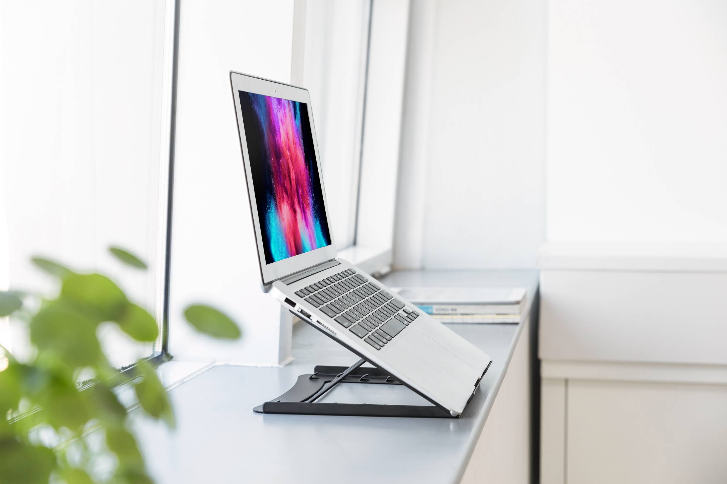 Laptop stojí na nastavitelném stojanu na okenním parapetu. Obrazovka zobrazuje pestré, rozmazané barvy.