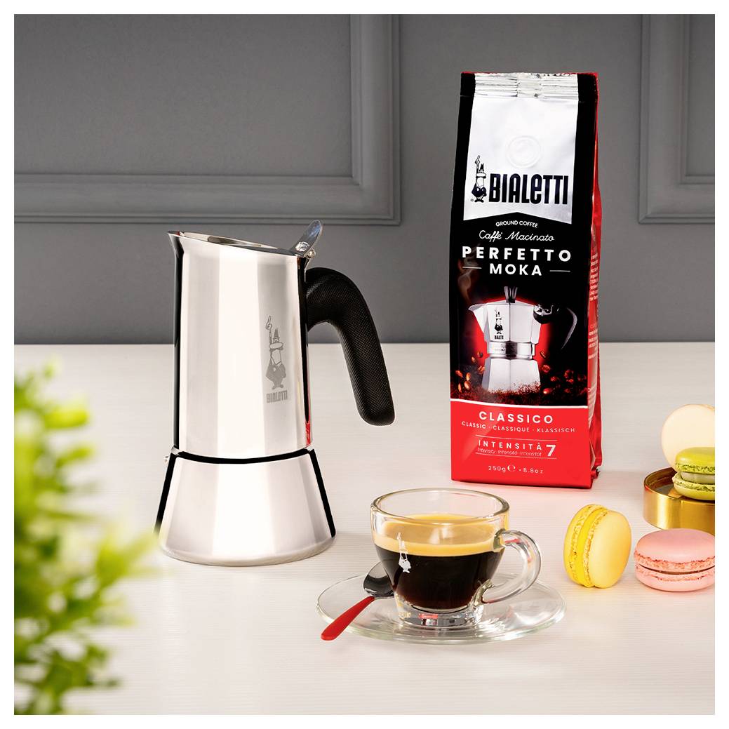 Kávový vařič na sporáku vedle balíčku kávy označeného "Bialetti Perfetto Moka", sklenice espresa a tři makronky na stole.