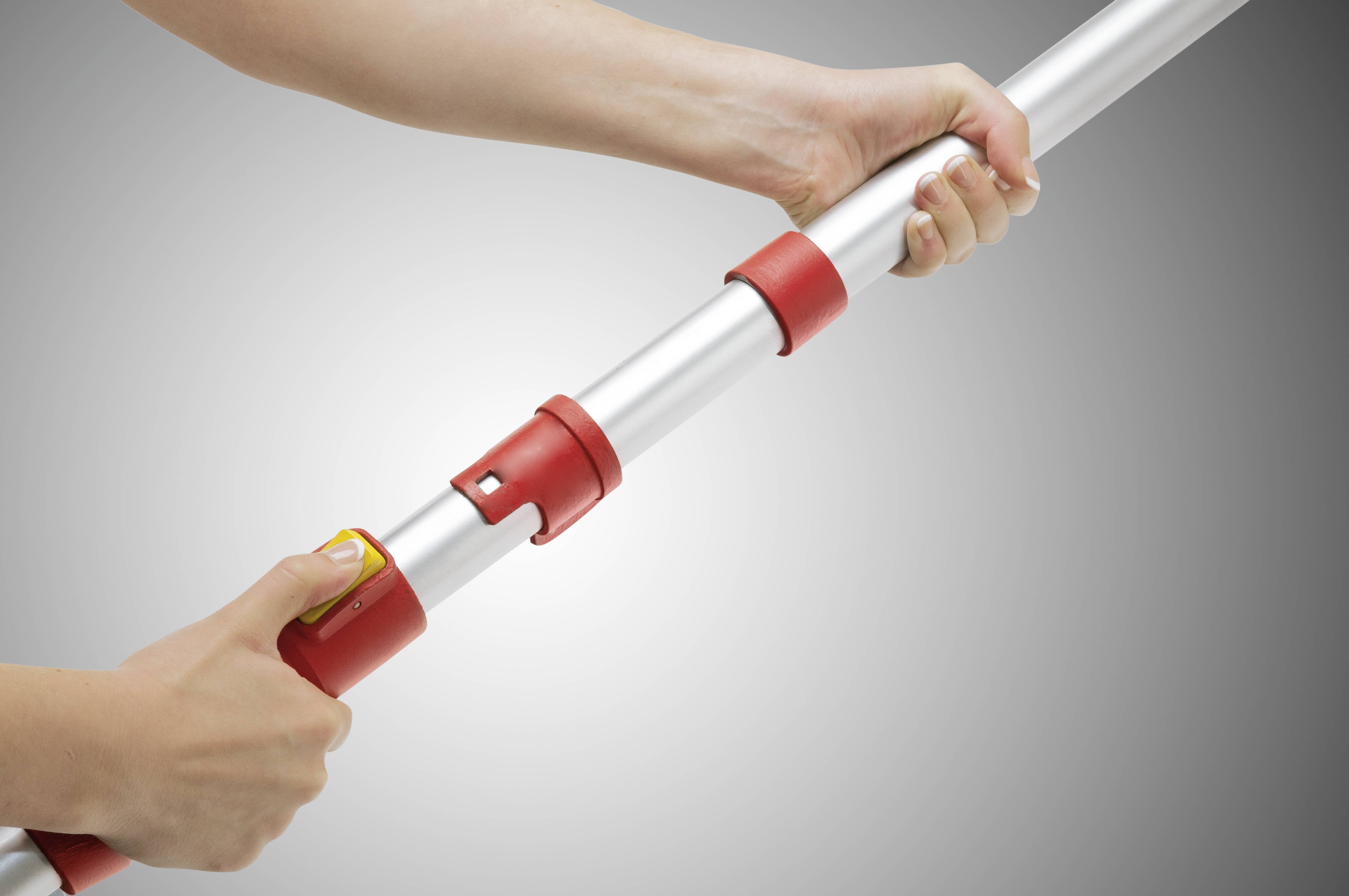 Osoba drží výsuvnou kovovou tyč s červenými spoji. Jedna ruka ovládá žluté tlačítko pro nastavení délky.